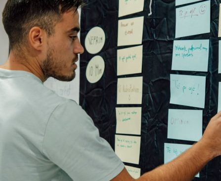 A man in a casual t-shirt organizes notes on a wall, focusing on a 'WORK OUT' sign.